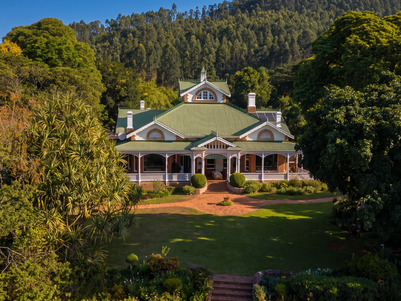 An aerial view of a charming, well-maintained heritage-style house featuring a green corrugated metal roof, wrap-around veranda, and lush, expansive landscaped gardens.