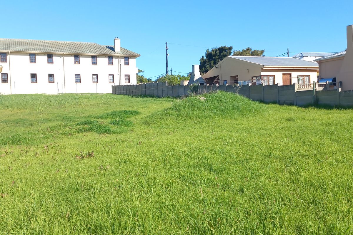 A view of a vacant residential plot featuring a grass lawn and concrete boundary wall, surrounded by neighboring residential buildings.