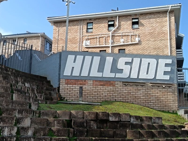 Exterior view of the Hillside apartment complex featuring a brick facade, security fencing, and a tiered retaining wall entrance.
