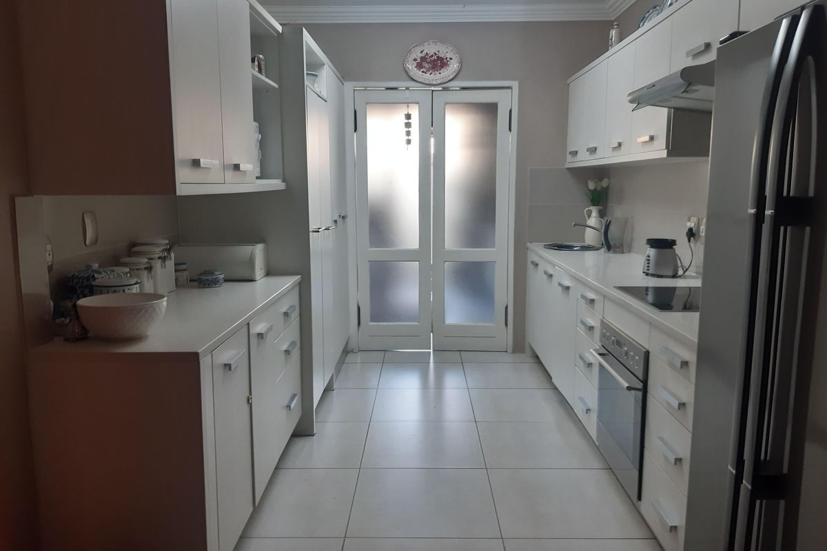 A well-maintained galley-style kitchen featuring white cabinetry, stone countertops, integrated oven, electric hob, and tiled flooring, leading to a glass-paneled exterior door.