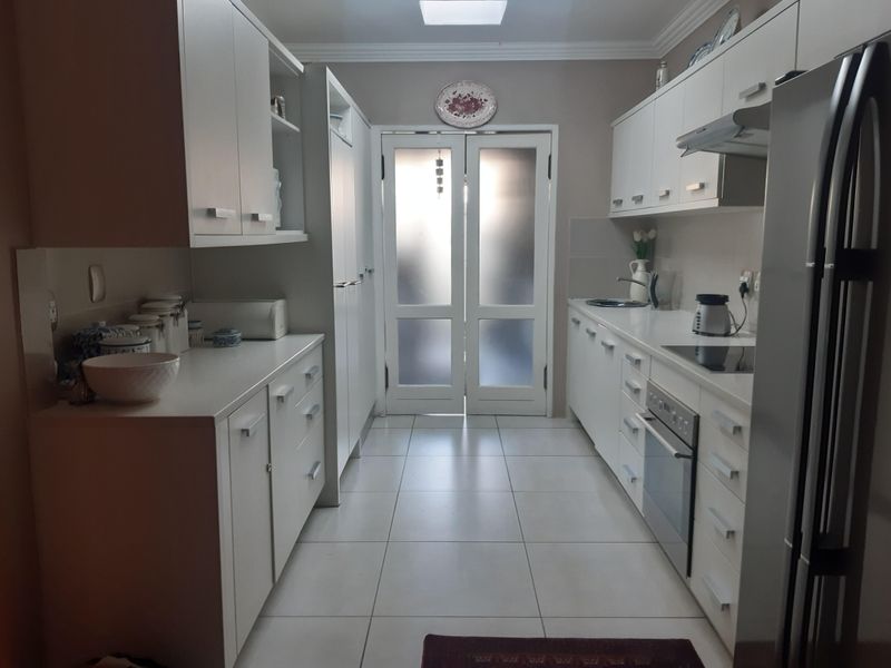 A well-maintained galley-style kitchen featuring white cabinetry, stone countertops, integrated oven, electric hob, and tiled flooring, leading to a glass-paneled exterior door.