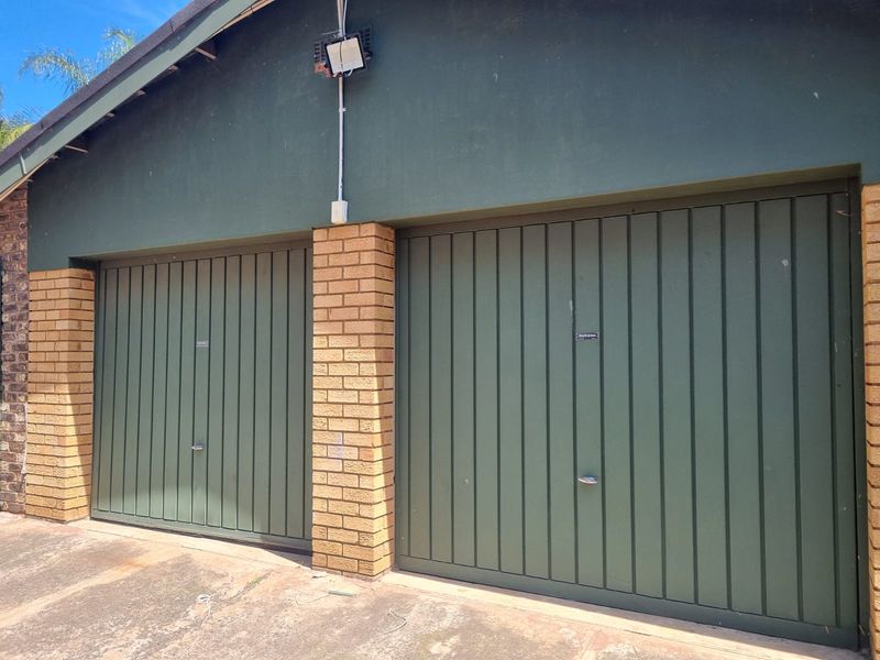 A double garage featuring green vertical-panel doors, brick support pillars, and an exterior floodlight, set above a concrete driveway.