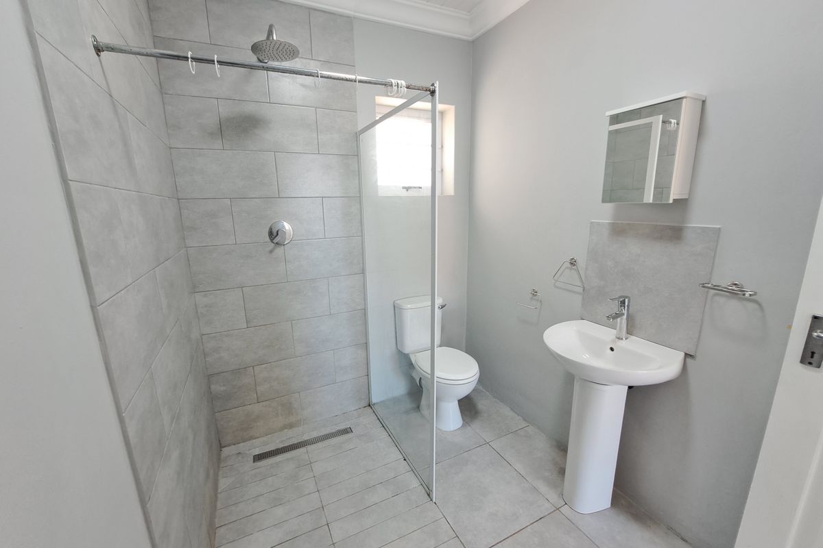 A modern, clean bathroom featuring a walk-in shower with a rain shower head, a pedestal basin, and a toilet, finished with neutral grey tiling.