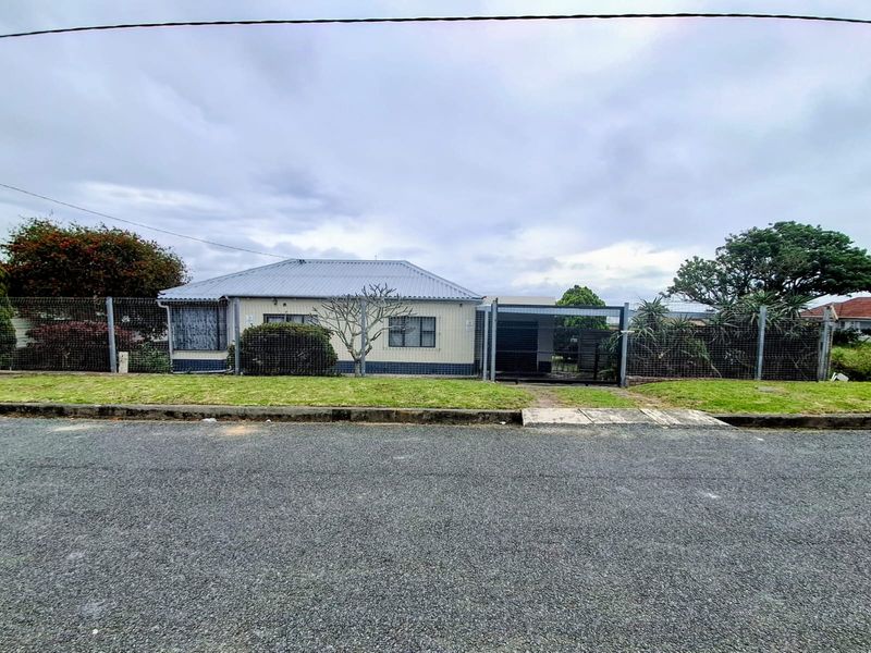 Front view of a single-story house featuring a corrugated metal roof, security fencing, and a paved driveway entrance.