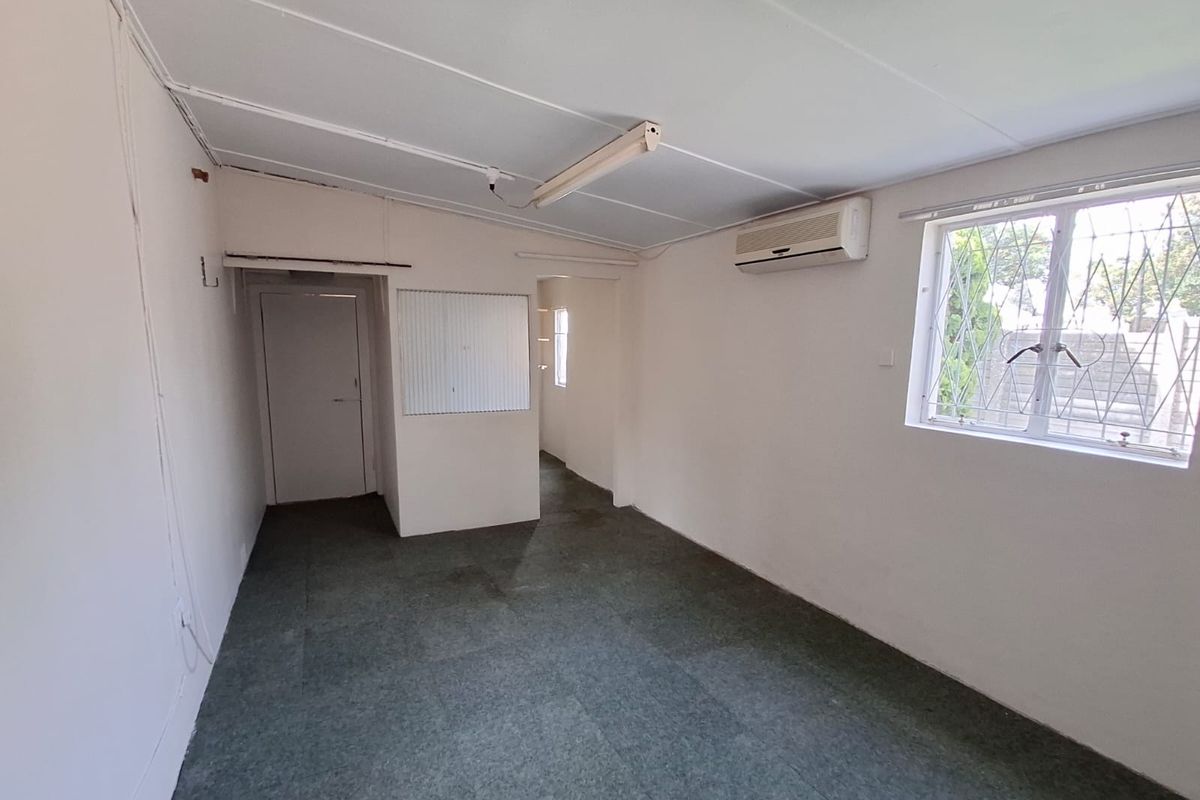 An empty, neutral-toned living area featuring a wall-mounted air conditioning unit, security bars on the window, and a simple ceiling light fixture.