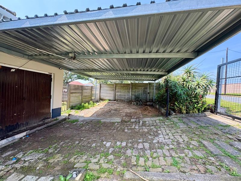 An open-sided carport featuring a corrugated metal roof, steel support structure, and cobblestone paving, enclosed by precast concrete boundary walls.