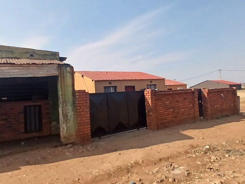 Front exterior view of a residential property featuring a brick boundary wall, metal gate, and corrugated metal roofing structure.