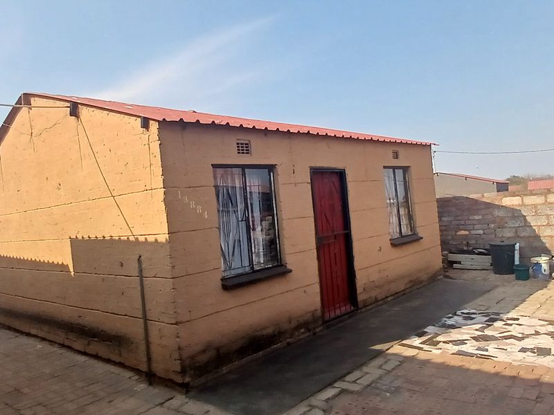 A modest single-story residential dwelling featuring a corrugated metal roof, security entrance gate, and paved exterior area with boundary walling.