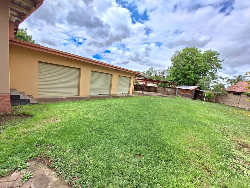 Spacious rear garden featuring a triple garage with roller shutter doors, a detached carport, and an outbuilding, set within a fenced property boundary.