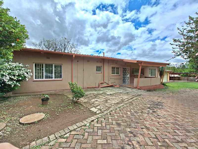 Rear view of the house featuring a paved driveway, covered patio area, and established garden surroundings with security bars on windows.