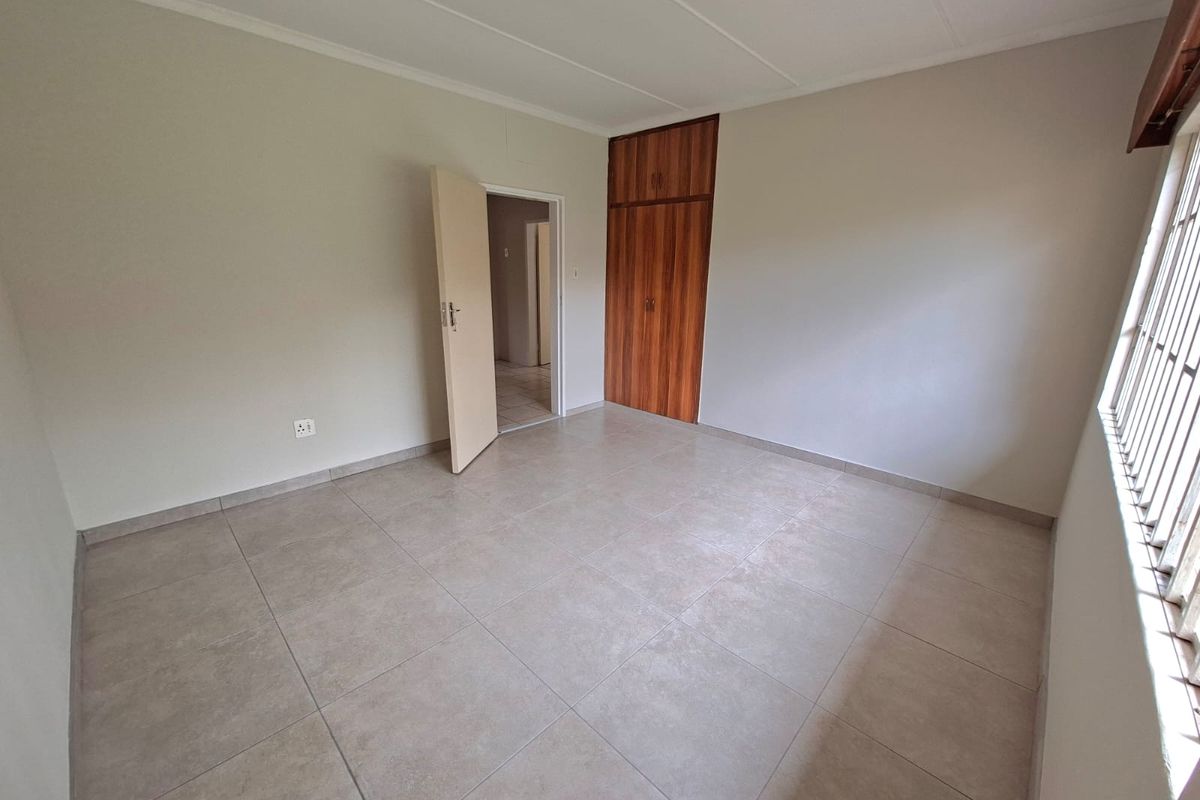 A clean, empty bedroom featuring neutral tiled flooring, built-in wooden cupboards, and security bars on the window.
