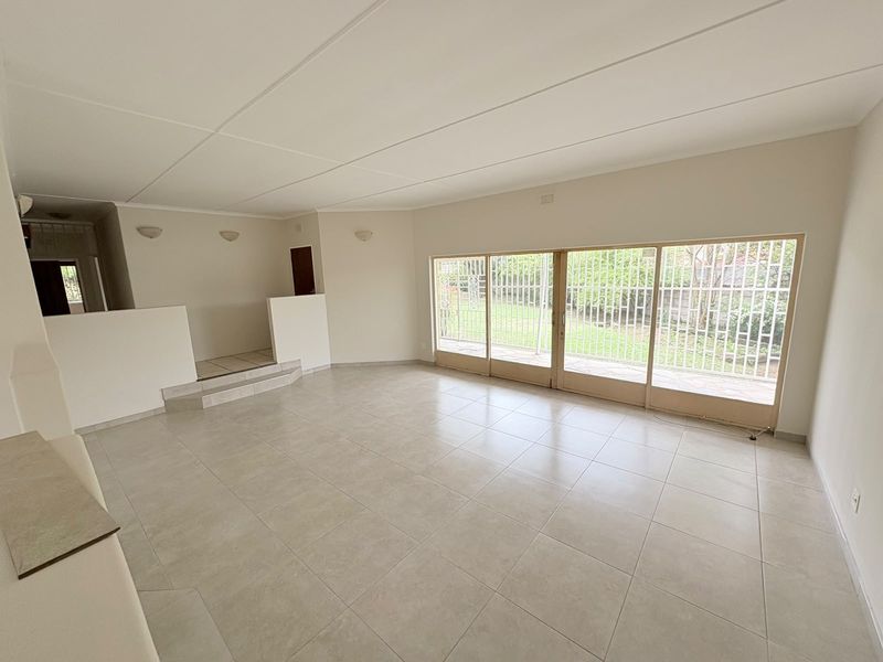 Spacious, light-filled living area featuring neutral tiled flooring, a raised platform section, and large sliding glass doors with security bars overlooking the garden.