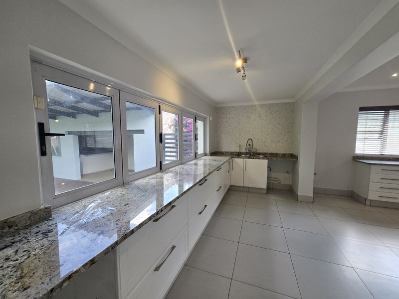 A bright, modern kitchen featuring granite countertops, ample built-in cabinetry, a double sink, and large windows overlooking the outdoor patio area.