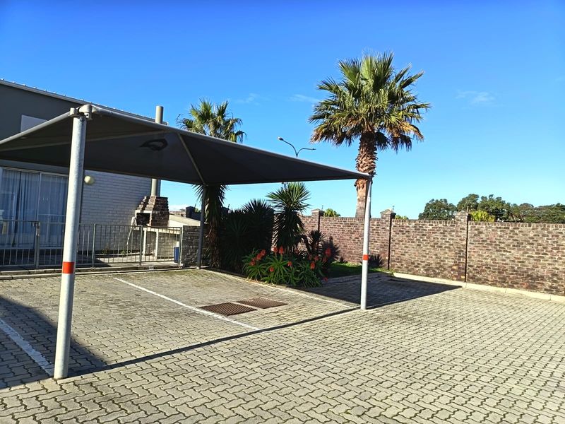 Exterior view of a paved parking area featuring a shade sail carport, brick boundary wall, and a built-in outdoor braai facility.