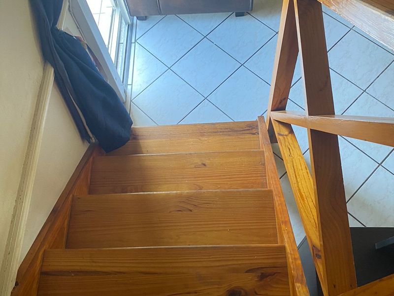 A view looking down a wooden staircase with a timber balustrade, leading to a tiled living area with access to a sliding glass door.