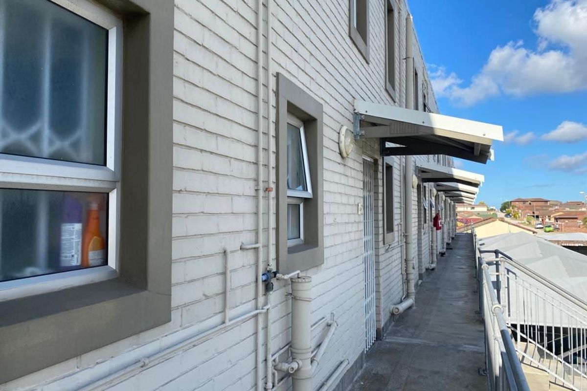 Exterior view of an apartment building walkway featuring painted brick walls, metal security gates, and protective awnings over the unit entrances.