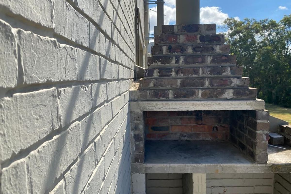 Outdoor patio area featuring a built-in brick braai structure against a painted brick wall, offering potential for an upgraded entertainment space.