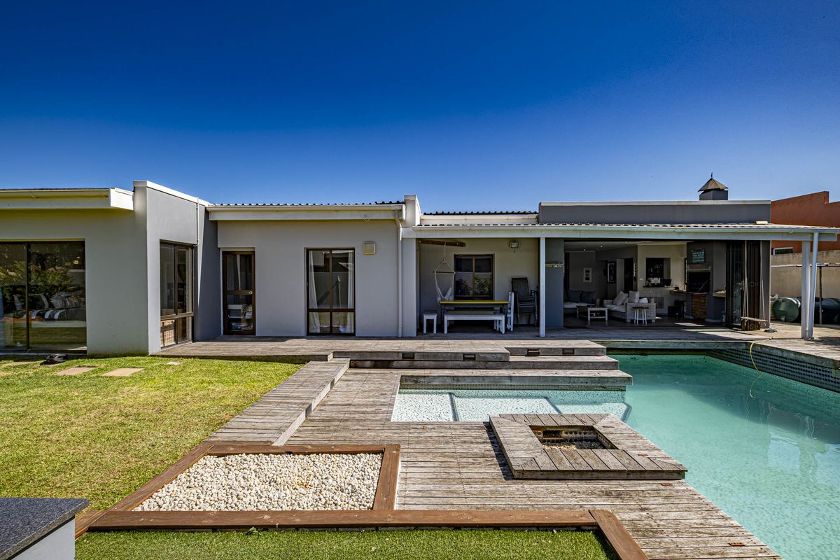 Rear exterior view featuring a swimming pool, extensive timber decking, a covered patio area with a built-in braai, and a well-maintained garden.