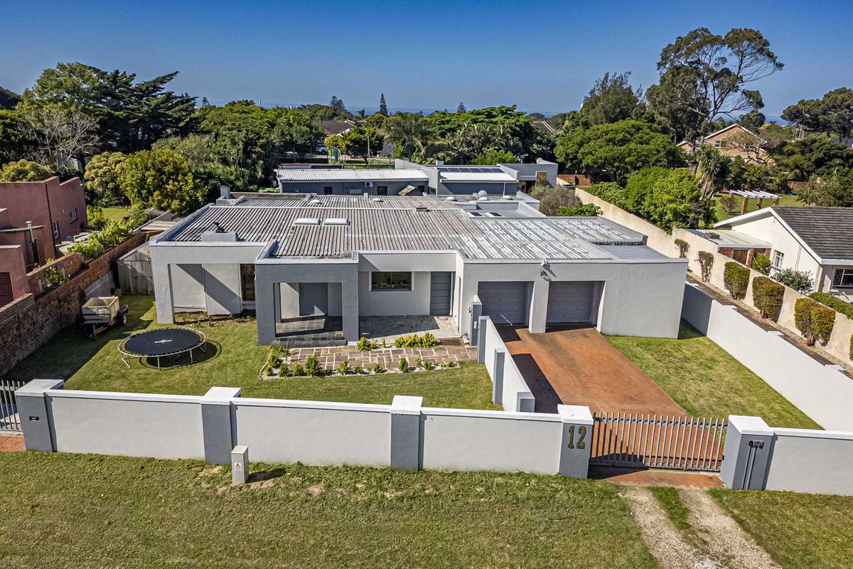 Aerial view of a modern single-story residence featuring a corrugated metal roof, double garage, paved driveway, and a secure perimeter wall with a sliding gate.