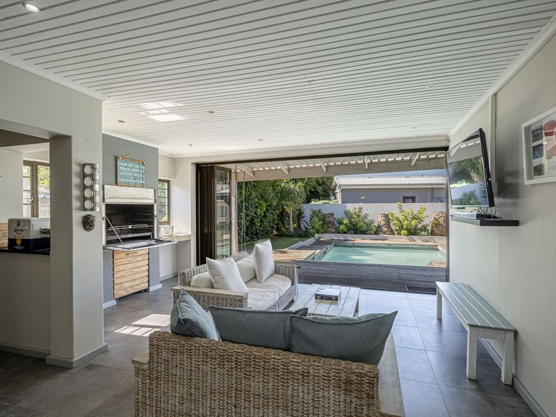 A spacious covered patio featuring a built-in braai, tiled flooring, and seamless flow through bi-fold doors to a wooden deck and swimming pool area.