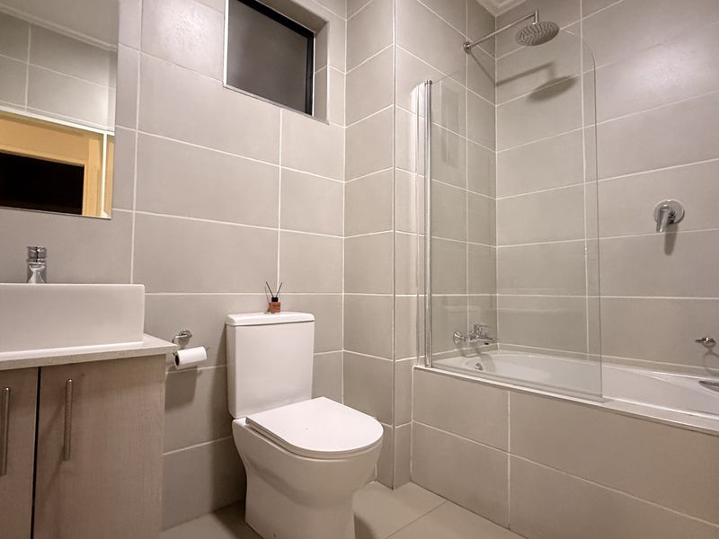 Modern bathroom featuring neutral wall and floor tiles, a vanity with vessel sink, a toilet, and a bathtub with a glass shower screen.