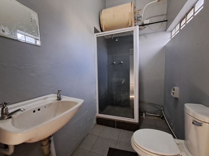 A dated bathroom featuring a shower enclosure, pedestal basin, and toilet, with exposed plumbing and visible wear on wall finishes.