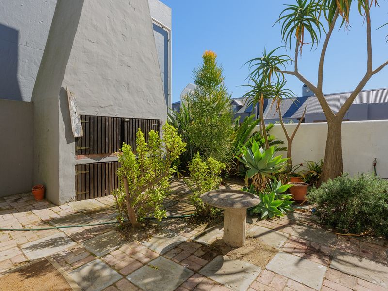 A private rear courtyard featuring a paved patio area and a built-in braai, surrounded by established garden landscaping and boundary walls.