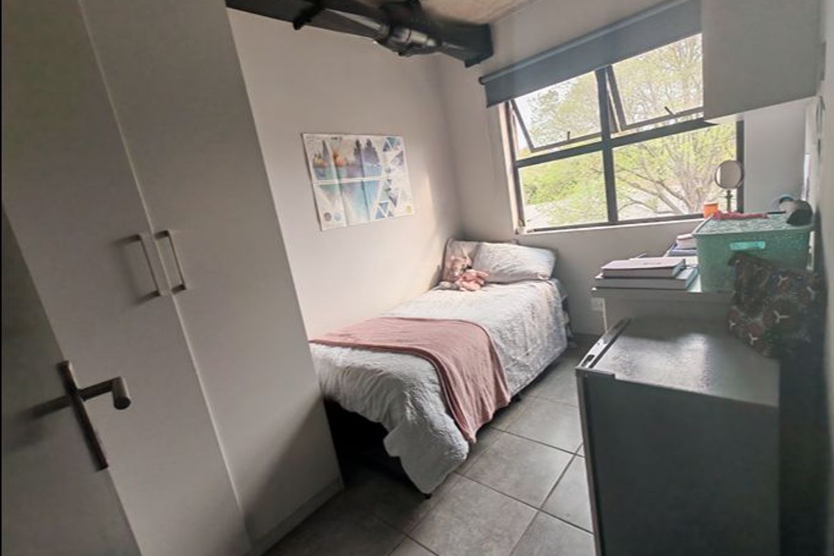 A compact bedroom featuring tiled flooring, a built-in wardrobe, and an exposed ceiling beam, with natural light provided by a casement window.