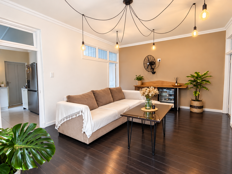 A well-maintained living room featuring dark timber flooring, decorative coving, and stylish pendant lighting, with an adjacent view into the kitchen area.