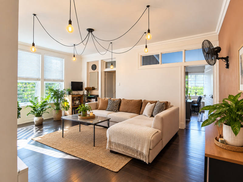 A bright and inviting living room featuring dark hardwood floors, crown molding, and a unique multi-pendant lighting fixture, with natural light from large windows.