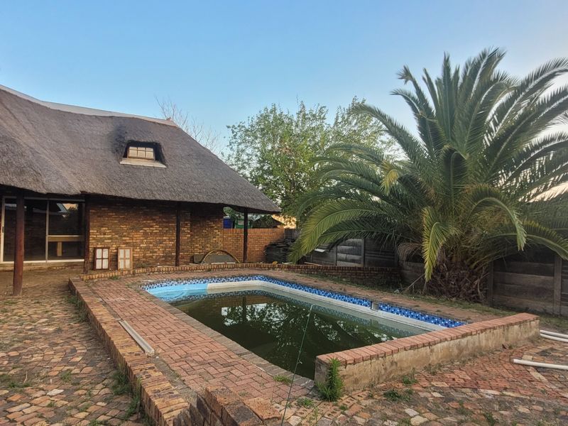 Rear exterior view featuring a swimming pool with brick surround, adjacent to a thatched-roof brick house with sliding door access.