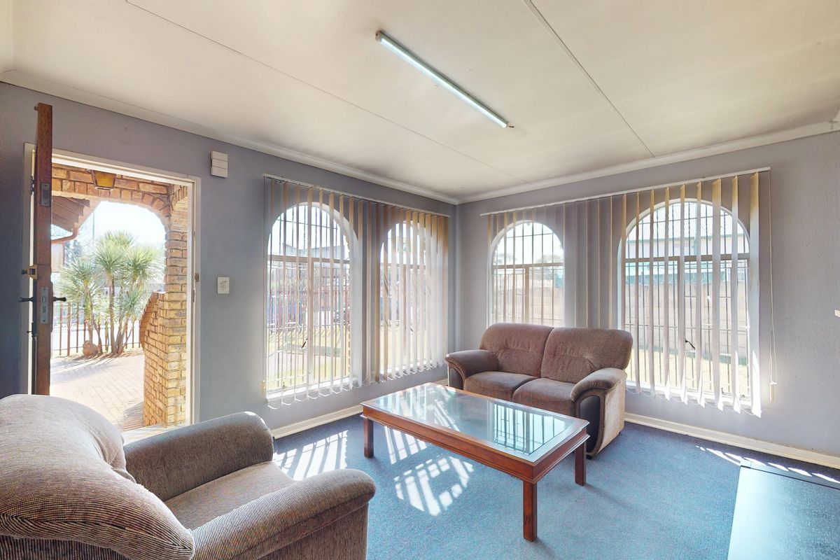 A living room featuring arched windows with vertical blinds, a brick feature wall, and carpet flooring, providing direct access to an outdoor area.