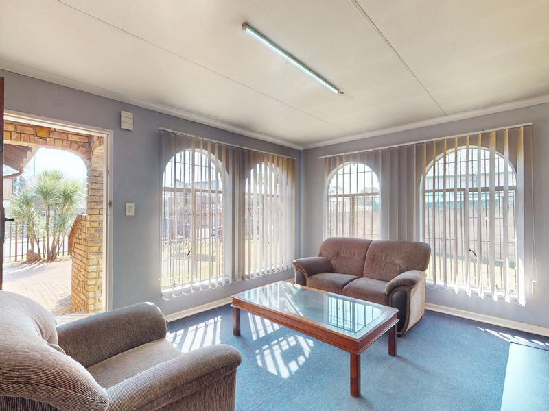 A living room featuring arched windows with vertical blinds, a brick feature wall, and carpet flooring, providing direct access to an outdoor area.