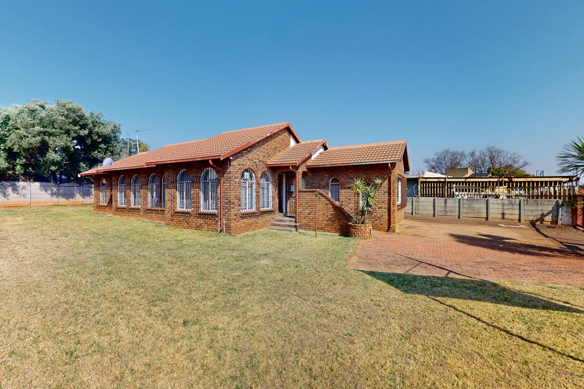 A rear view of a face brick house featuring arched windows with security bars, a tiled roof, and a spacious lawn with a paved driveway area.
