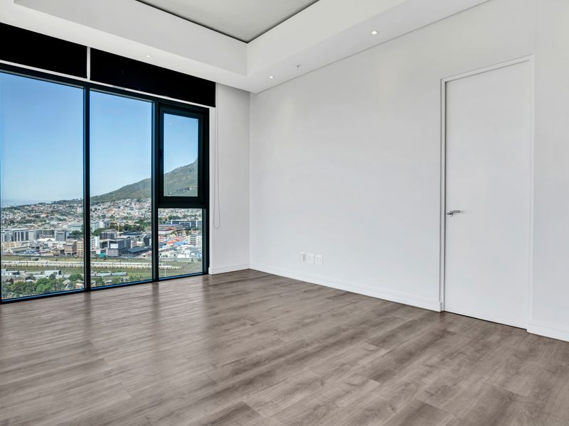 A bright, modern, and empty bedroom featuring floor-to-ceiling windows with expansive city and mountain views, laminate flooring, and recessed ceiling lighting.