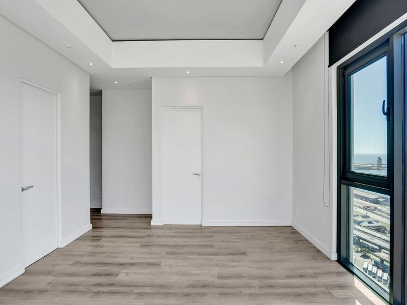 A bright, modern, and empty bedroom featuring high-quality laminate flooring, recessed ceiling detail, and a large window offering expansive coastal and city views.