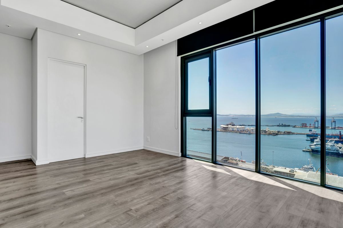 A bright, empty bedroom featuring modern laminate flooring, recessed ceiling lighting, and expansive floor-to-ceiling windows offering panoramic harbor and ocean views.