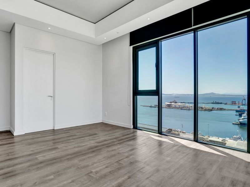 A bright, empty bedroom featuring modern laminate flooring, recessed ceiling lighting, and expansive floor-to-ceiling windows offering panoramic harbor and ocean views.