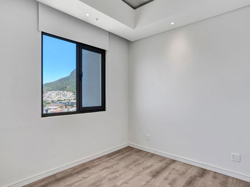 A bright, empty bedroom featuring modern laminate flooring, recessed ceiling lighting, and a window offering scenic mountain and city views.