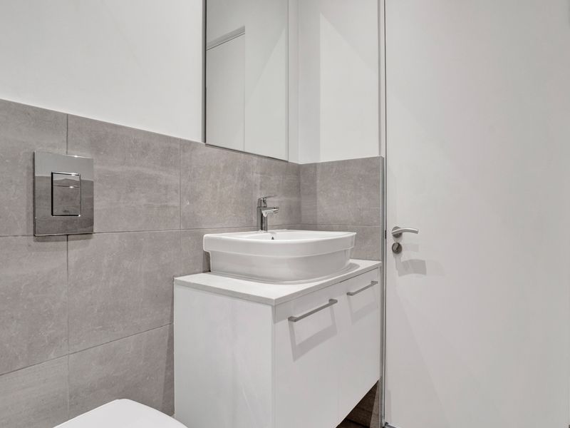 Modern bathroom featuring a sleek vanity cabinet with a vessel sink, wall-mounted toilet flush plate, and neutral grey wall tiling.