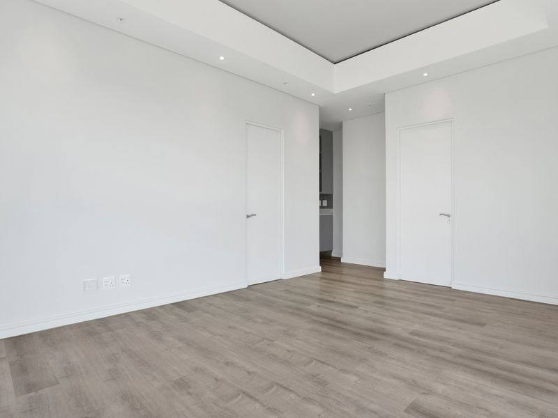 A bright, empty living area featuring modern laminate flooring, recessed ceiling lighting, and clean white walls with flush panel doors.