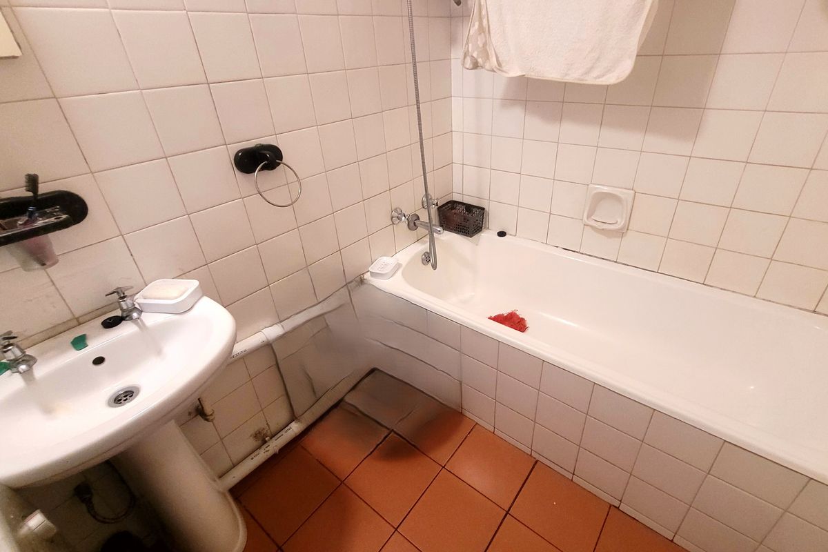 A functional bathroom featuring a pedestal washbasin and a bathtub with shower mixer, finished with white wall tiles and terracotta-style floor tiles.