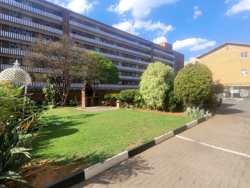 A well-maintained communal garden area featuring a brick built-in braai, manicured lawn, and paved driveway access within the apartment complex grounds.
