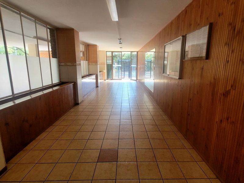 A spacious, dated communal hallway featuring timber wall paneling, tiled flooring, and large glass partitions leading to the building entrance.