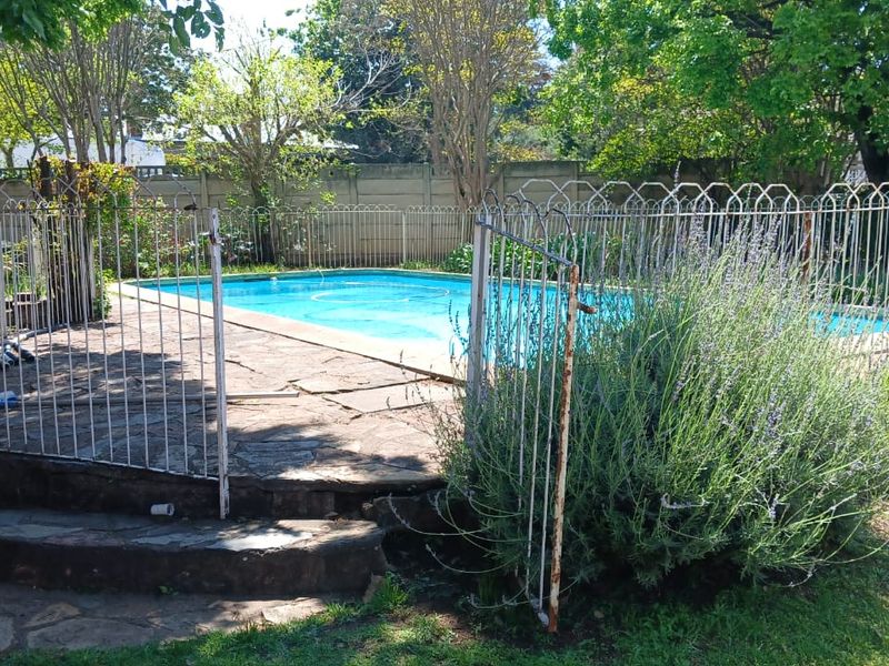 Outdoor swimming pool area featuring stone paving and metal safety fencing, surrounded by established garden greenery and a concrete boundary wall.