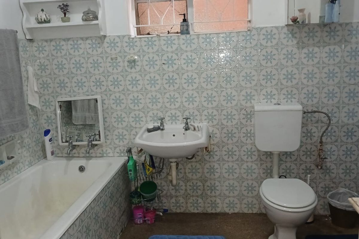 A dated bathroom featuring a wall-mounted basin, built-in bathtub, and toilet, finished with patterned wall tiles and a window with security bars.