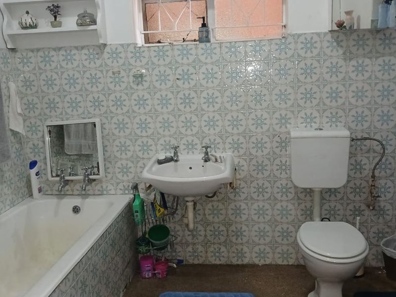 A dated bathroom featuring a wall-mounted basin, built-in bathtub, and toilet, finished with patterned wall tiles and a window with security bars.