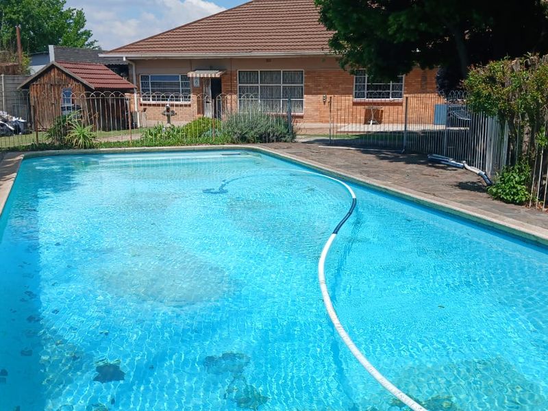 A backyard view featuring a swimming pool with surrounding paving, metal fencing, and a brick house exterior with a tiled roof.