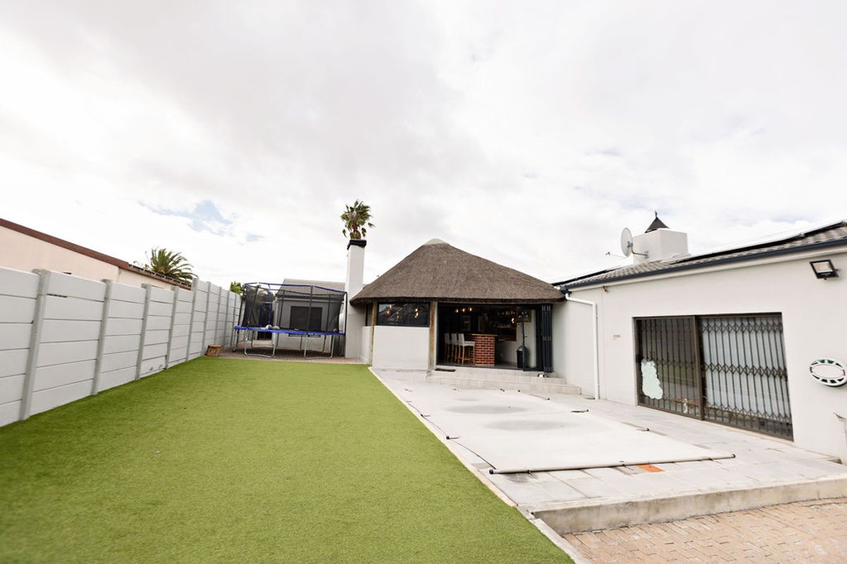 Rear garden area featuring an artificial lawn, paved patio with a pool cover, and a thatched-roof entertainment structure.