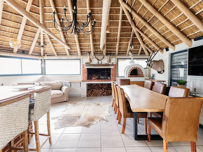 Spacious open-plan living and dining area featuring a high thatched roof with exposed timber rafters, a brick fireplace, and a built-in pizza oven.